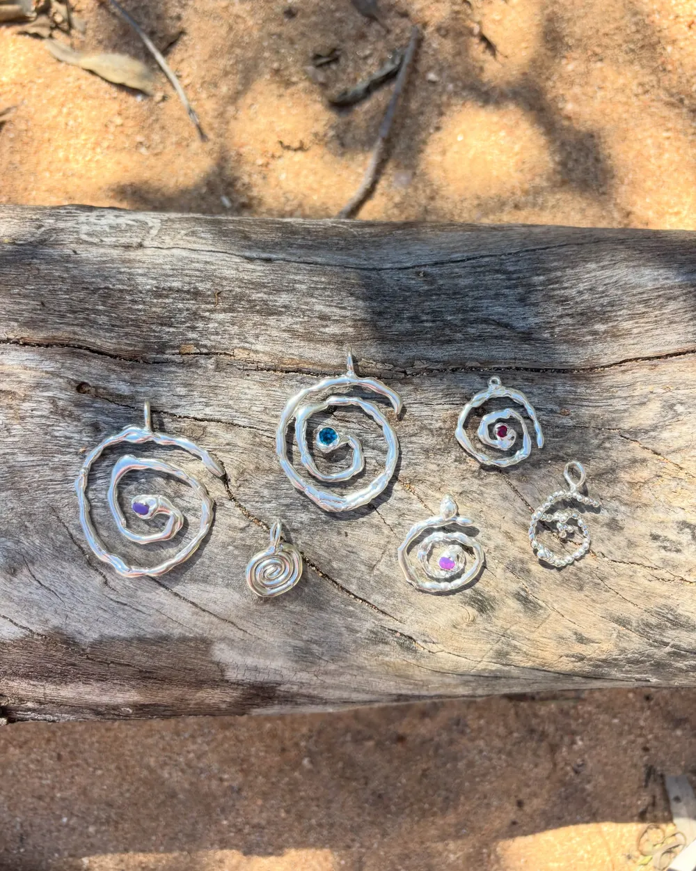 Six handmade silver spiral pendants with coloured stones laid out on weathered wood